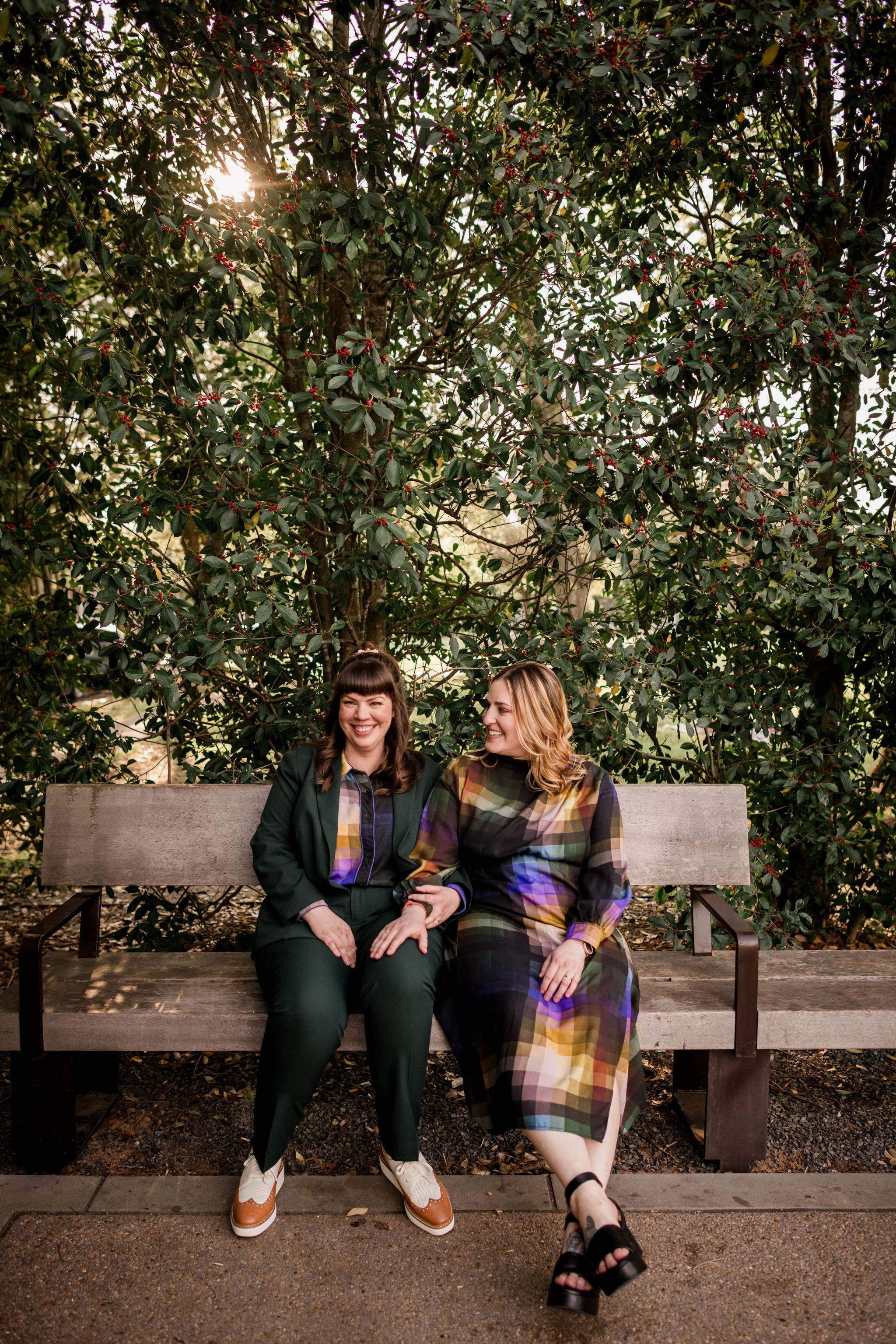 Same sex couple smiling at each other holding a pink and peach colored bouquet, one with light brown hair wearing a mustard jacket and white shirt and the other with raven colored hair wearing a cranberry colored jacket over a white wedding gown photo by Wedding Photographer in Houston, Jamie Hardin Photography